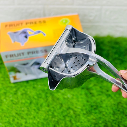 Metallic fruit press held by a hand on a green surface with a product packaging in the background.