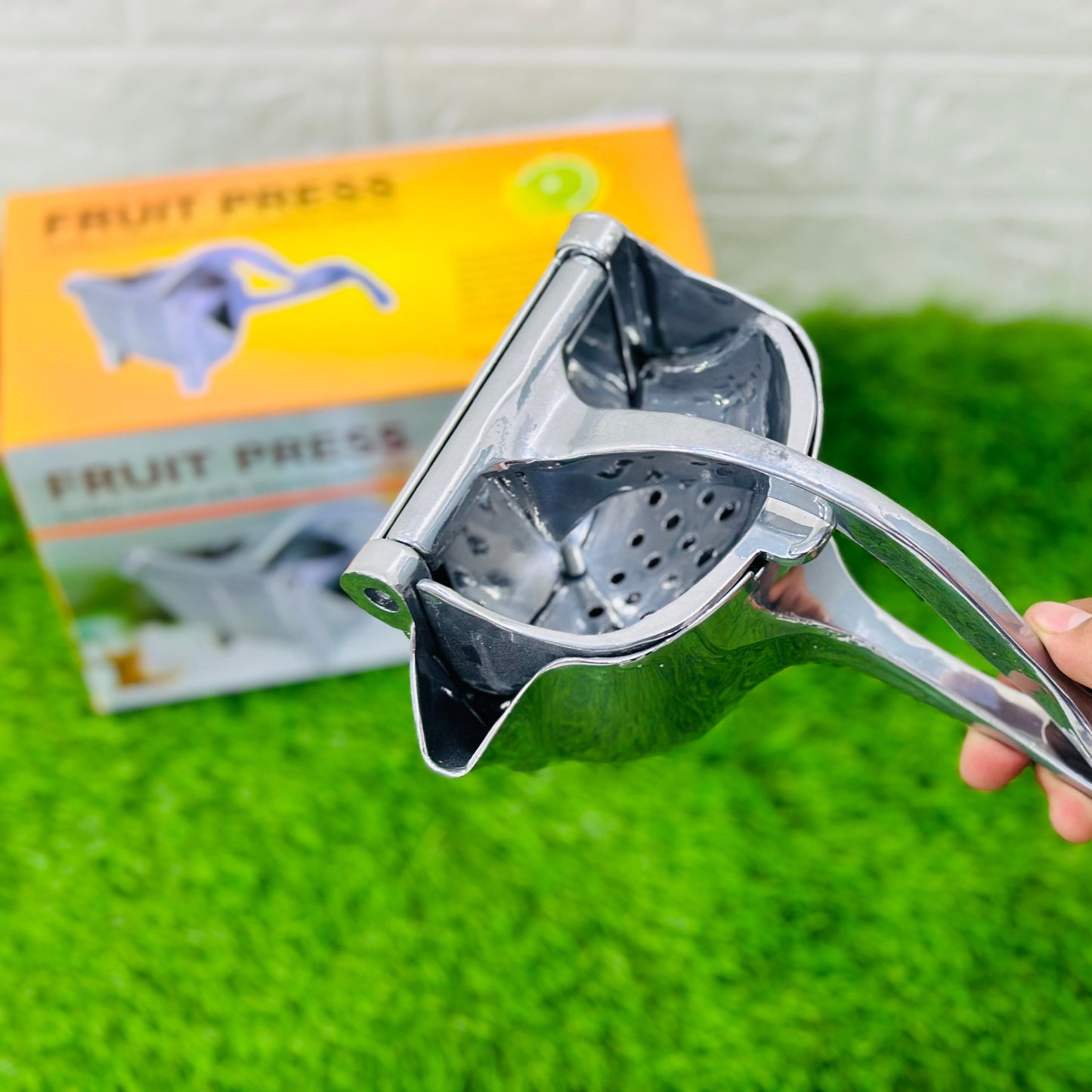 Metallic fruit press held by a hand on a green surface with a product packaging in the background.