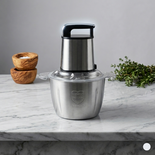 Stainless steel food chopper on a marble countertop with wooden bowls and herbs in the background.