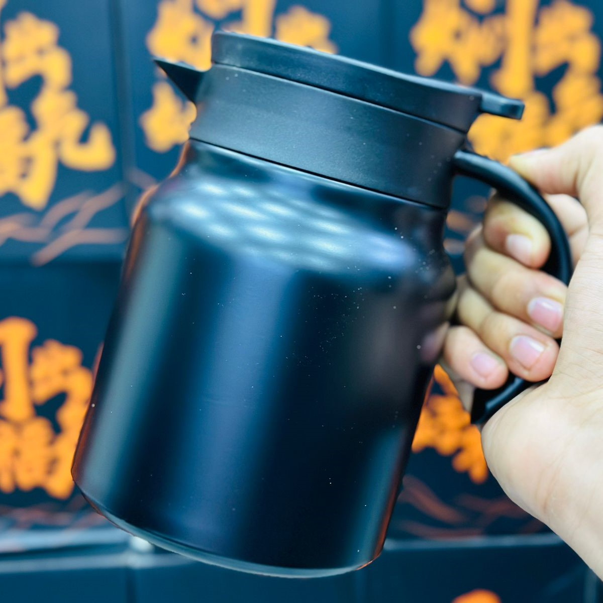 A hand holding a black stainless steel thermos with a lid and a handle, against an oriental-inspired background.