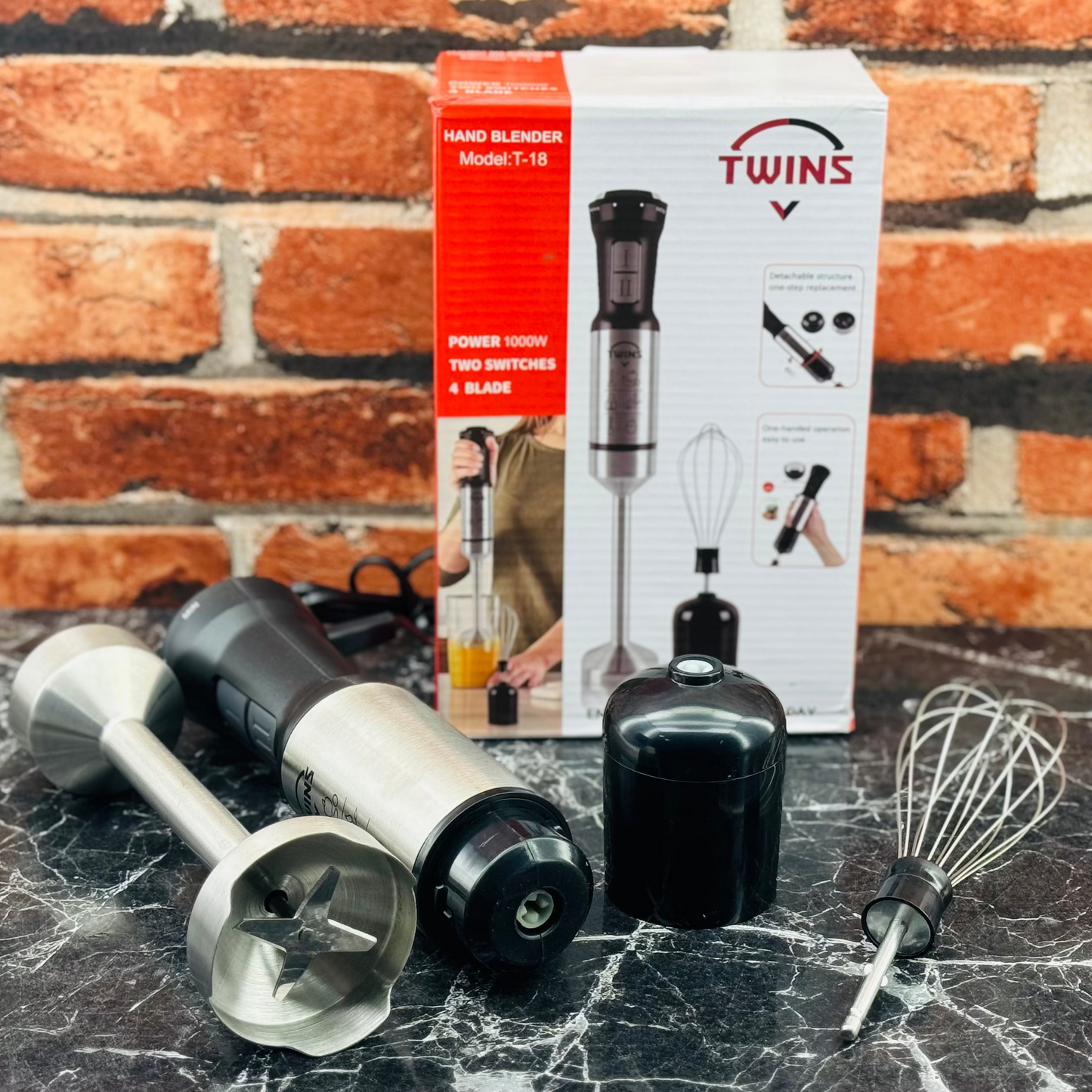 A hand blender set by Twins, including a blender, a whisk, and two stainless steel attachments, displayed next to its packaging box on a marble surface.