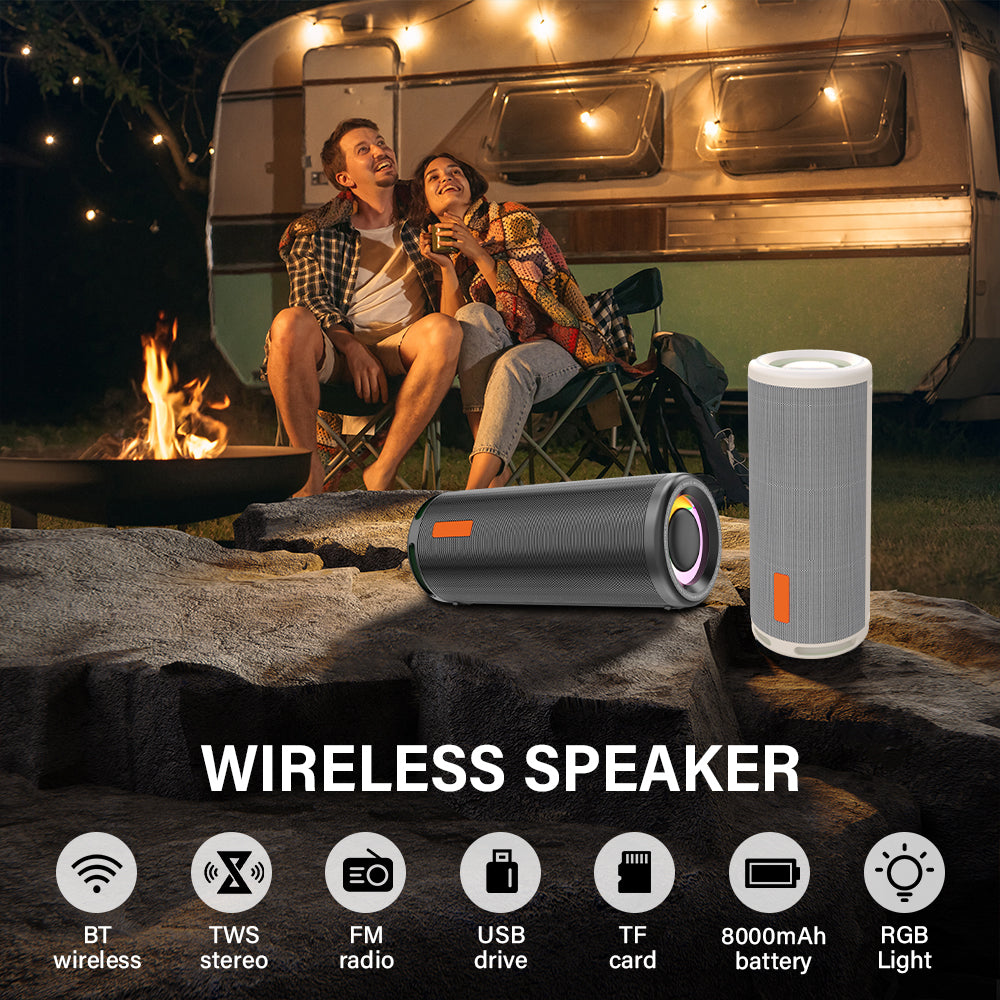 Two people sitting by a campfire with a wireless speaker and a portable device on a rock, with a camper in the background.