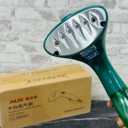 A handheld garment steamer in green color, with a large water tank and a handle, displayed in front of a cardboard box with text on it.