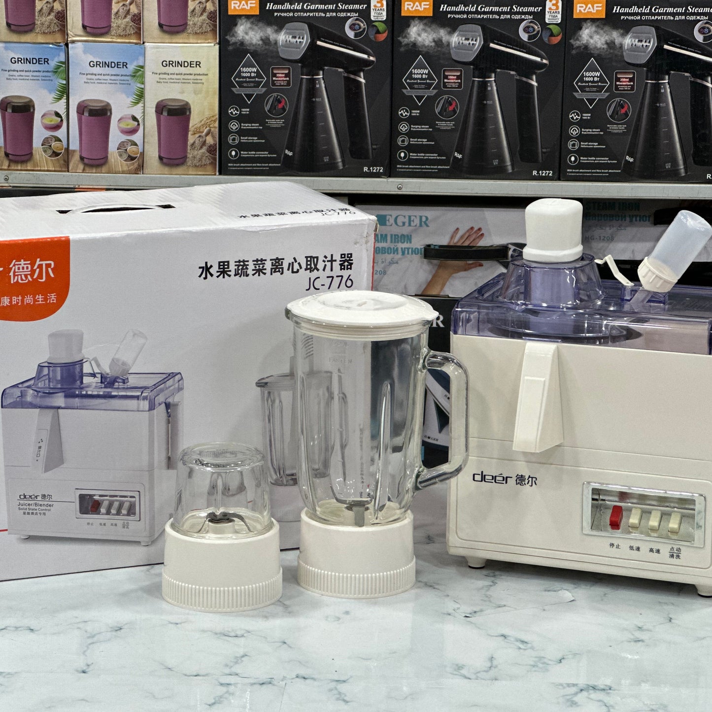 A white Deer brand juicer blender set, including a juicer, blender jug, and grinder, displayed on a counter with its packaging box in the background.