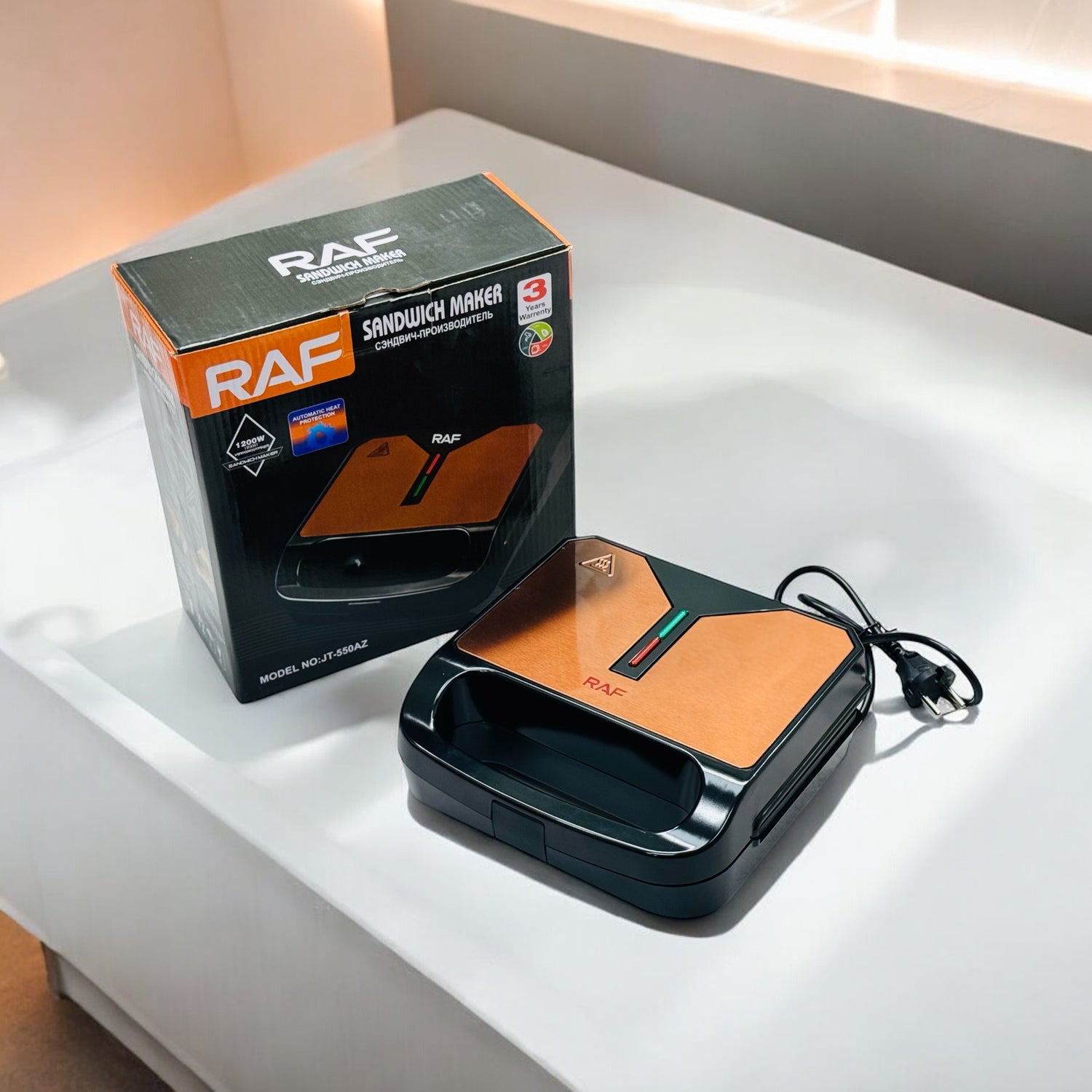 A Raf Sandwich Maker 1200W with its packaging box. The maker is primarily black and orange, with a digital display and control buttons visible.