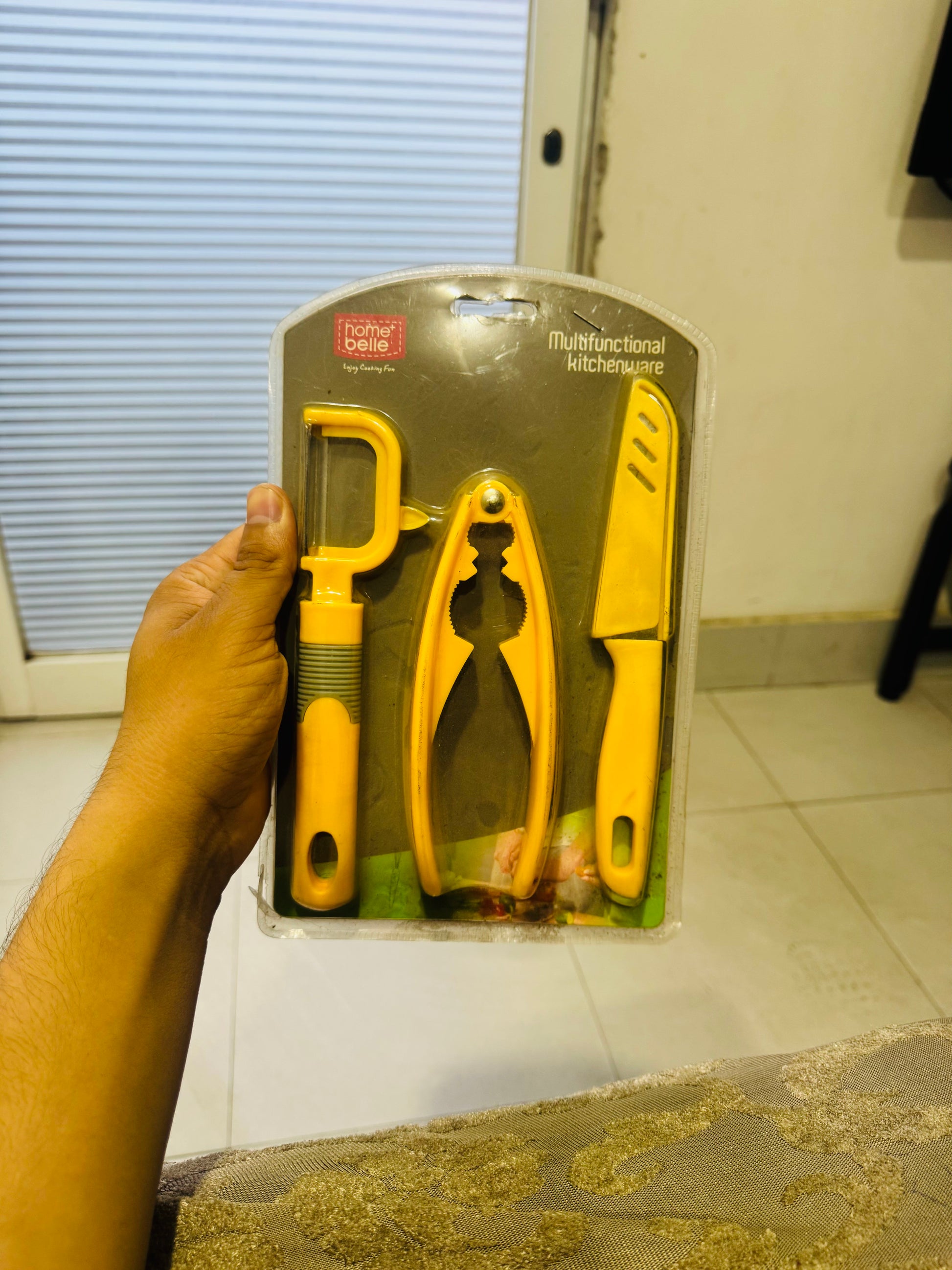 Set of yellow kitchen tools in packaging held by a hand on a tiled floor.