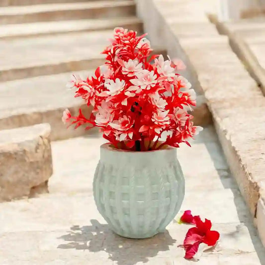 A artificial flora piece with a green pot, containing red and white artificial flowers, placed on a set of stairs with a pink flower petal scattered beside it.