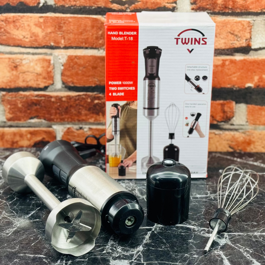 A hand blender set by Twins, including a blender, a whisk, and two stainless steel attachments, displayed next to its packaging box on a marble surface.