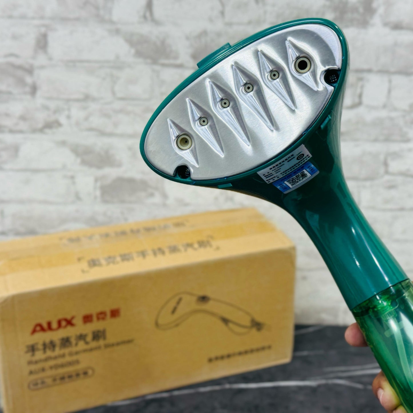 A handheld garment steamer in green color, with a large water tank and a handle, displayed in front of a cardboard box with text on it.